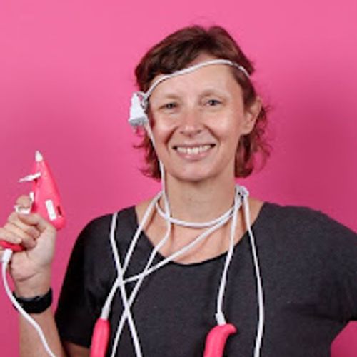 Smiling woman with short curly hair wearing a dark t-shirt, holding a pink hot glue gun and draped in cords with several other glue guns, standing against a bright pink background.